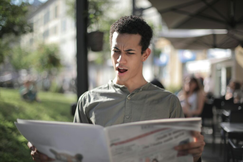 A young man is surprised while reading a newspaper at an outdoor cafe.