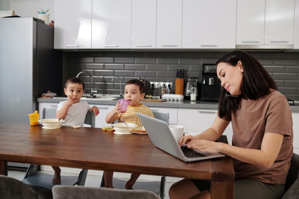 A mother multitasking on a laptop while her two young sons enjoy breakfast in the kitchen.
